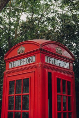 Londra, İngiltere - 20 Haziran 2020: Ağaçlara karşı kırmızı telefon kulübesinin alçak açılı görüntüsü. Kırmızı telefon kulübeleri dünyadaki mevcut veya eski İngiliz kolonilerinde bulunabilir..