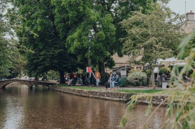 Bourton-on-the-Water, İngiltere - 10 Temmuz 2020: İngiltere 'nin Cotswolds kırsalındaki ünlü bir köy olan Bourton-on-the-Water' daki River Windrush 'da dinlenen insanlar. Seçici odak.