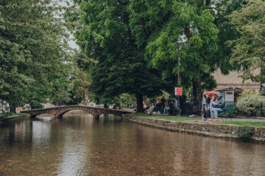Bourton-on-the-Water, İngiltere - 10 Temmuz 2020: İngiltere 'nin Cotswolds kırsalındaki ünlü bir köy olan Bourton-on-the-Water' daki River Windrush 'da dinlenen insanlar. Seçici odak.