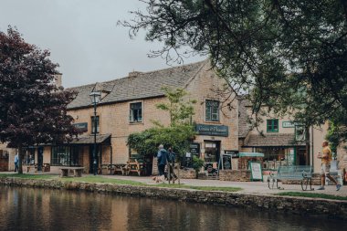 Bourton-on-the-Water, İngiltere - 10 Temmuz 2020: İnsanlar İngiltere 'nin Cotswolds kırsalındaki ünlü bir köy olan Bourton-on-the-Water' daki River Windrush nehri boyunca kafelerin ve restoranların önünden geçiyor.