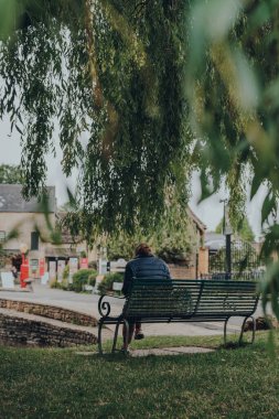 Bourton-on-the-Water, İngiltere - 12 Temmuz 2020: İngiltere 'nin Cotswolds kırsalındaki ünlü bir köy olan Bourton-on-the-Water' da, River Windrush 'un yanındaki bir ağacın altında bir kadının arka görüntüsü. Seçici odak.