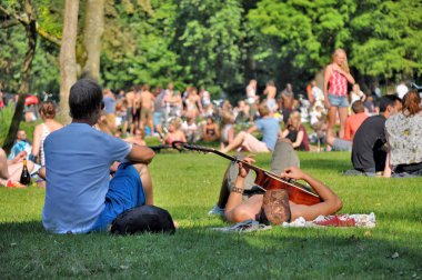 Amsterdam, Netherlands - Juli 23, 2012: Two young men enjoy a summerday in the Vondelpark in Amsterdam. One of them is playing guitar, while the other one is looking at the people on the meadows in the background.