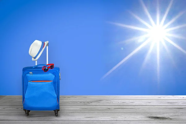 Blue suitcase with summer hat and fashionable sunglasses on shining fair bright sun with beams at clear blue sky background and wooden floor, summer vacation travel concept, free space with a place for copying