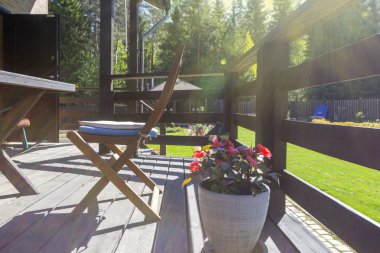 Kır evinin modern arka bahçesinde doğal ahşap açık rahat teras. Güneşli sabah, bahçe arka planda tencere çiçekler.