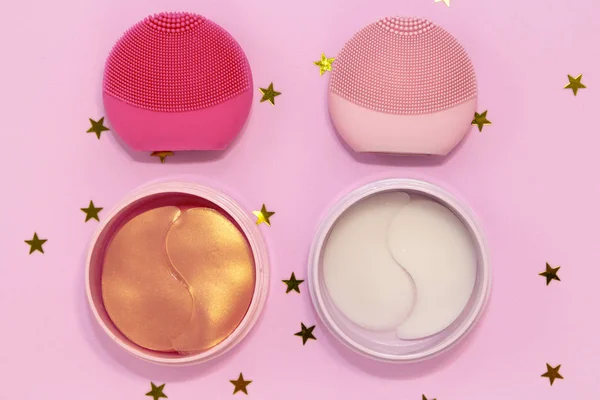 Silicone facial cleansing brushes with cleansing brush for massaging skin care and hydrogel cosmetic eye patch on pink background with golden stars. Flat lay, top view.