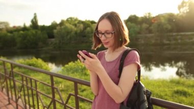 Smartphone ayakta açık havada köprüde kullanan öğrenci kız. Bir telefon, Internet tarama hipster manifatura ve iletişim. Günbatımı.