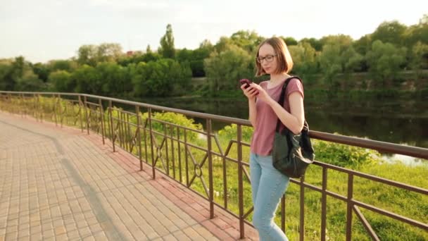 Trendy kız bardaklarda Avrupa şehirde smartphone kullanarak. Orada açık havada köprüde ayakta. Günbatımı. — Stok video