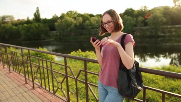 Smiling girl in glasses using smartphone standing outdoors on the bridge. — Stock Video