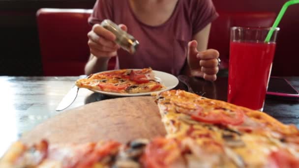 Casual menina pimentas um pedaço de pizza, come com um garfo e faca, sentado em um restaurante . — Vídeo de Stock