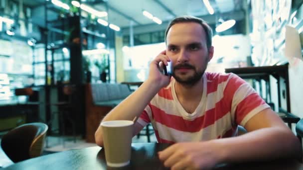 Un joven barbudo hablando por teléfono sentado en un café de la ciudad . — Vídeos de Stock