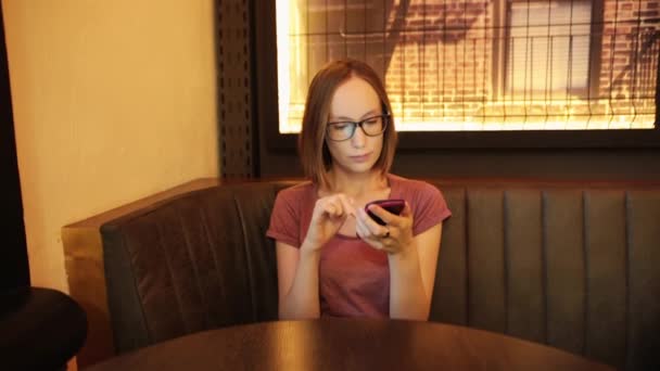 Mensaje de texto de mujer joven positiva en el teléfono inteligente moderno en la cafetería — Vídeos de Stock