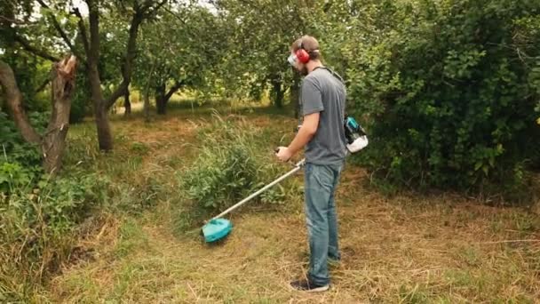 Homem barbudo jovem cortando grama verde no pomar com cortador de cordas . — Vídeo de Stock