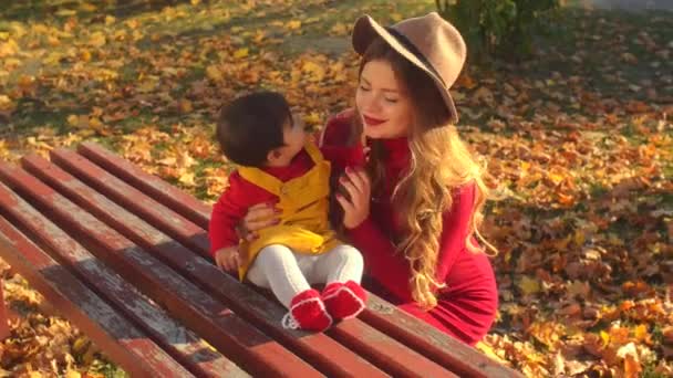 Giovane madre alla moda e bambino multietnico in abiti luminosi che giocano fuori sulla panchina . — Video Stock