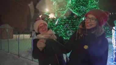 Anne ve kızı maytap kar yağışı altında dans sokakta Noel ağaç yakın:.
