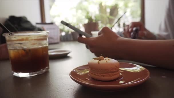 Menina usa um smartphone enquanto está sentado em um café e come macaron francês . — Vídeo de Stock