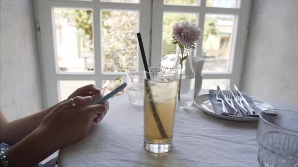 Ein junges Mädchen benutzt ein Smartphone in einem Café neben ihrer Holunderlimonade. — Stockvideo