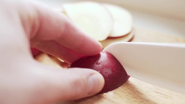 Großaufnahme von Händen, die einen roten Apfel mit einem weißen Keramikmesser auf dem Küchentisch schneiden. — Stockvideo