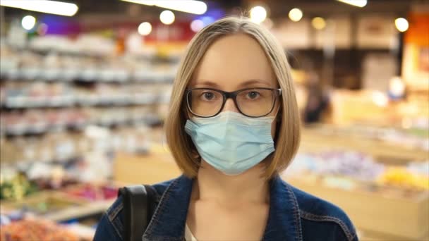 Porträtt av en ung kaukasiska flicka i en skyddande medicinsk mask och glasögon. slow motion i snabbköpet, närbild. — Stockvideo