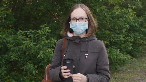 Retrato de una joven con una máscara médica con café para llevar en las manos, al aire libre . — Vídeos de Stock