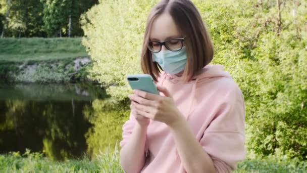 Дівчина в медичній масці, окулярах, рожевому светрі і короткій стрижці за допомогою смартфона . — стокове відео