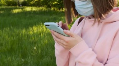 Koruyucu tıbbi maske takan ve pembe kapüşonlu gözlüklü bir kız akıllı telefon kullanıyor ve kuryeye mesaj yazıyor..