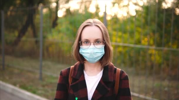 Une fille caucasienne en lunettes, masque médical de protection et un manteau regarde attentivement la caméra. — Video