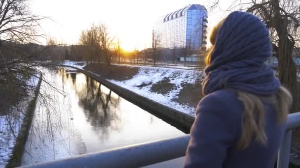Hermosa chica de pie en el puente y pensando a lo largo del muelle en el puerto, y viendo la puesta de sol, el cielo se refleja en el agua, hielo de primavera que se derrite en el agua. 4k — Vídeos de Stock