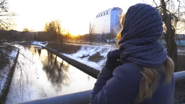 Όμορφο κορίτσι στέκεται στη γέφυρα και σκέφτεται κατά μήκος της προβλήτα στο λιμάνι, και βλέποντας το ηλιοβασίλεμα, ο ουρανός αντανακλάται στο νερό, άνοιξη πάγος λιώνει στο νερό. 4K — Αρχείο Βίντεο