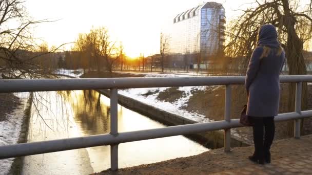 Vacker flicka som står på bron och tänker längs piren i hamnen, och titta på solnedgången, himlen reflekteras i vattnet, vårisen smälter på vattnet. 4K — Stockvideo