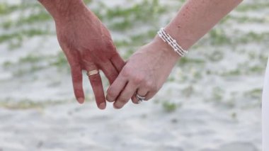 Bir kadının eli, bir erkeğin elini, arka plandaki kar beyazı çimenli kuma doğru tutar. Mücevher ve alyanslar. Genç adam kıyıda duruyor ve birlikte 4 bin dolar harcıyor.