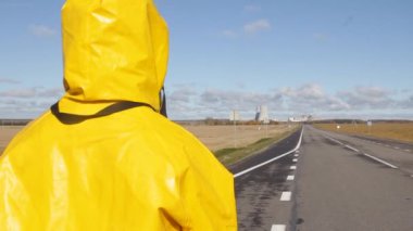 Sarı elbiseli bir adam biyolojik silahlardan ve radyasyondan korunuyor asfaltın yanındaki sokakta duruyor ve özel ekipmanlar kullanıyor ve açık bir güneşli bir günde nükleer santrale bakıyor.