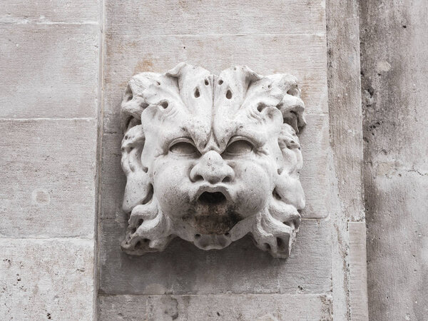 Closeup photograph of an old historic fountain squinting caricature face with large cheeks and open mouth protruding from a white masonry stone wall in Dubrovnik Croatia.