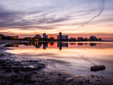 Güzel renkli gün batımı turuncu, pembe, mavi ve mor bulutlar gökyüzünde yukarıdaki ve Montrose Beach dalgaların karaya attığı odun ön planda bir parçası ile yağmur su birikintileri yansıtan ufukta binalar ile.