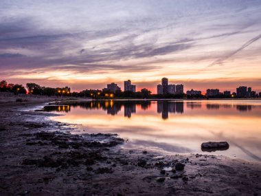 Güzel renkli gün batımı turuncu, pembe, mavi ve mor bulutlar gökyüzünde yukarıdaki ve Montrose Beach dalgaların karaya attığı odun ön planda bir parçası ile yağmur su birikintileri yansıtan ufukta binalar ile.
