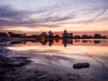 Güzel renkli gün batımı turuncu, pembe, mavi ve mor bulutlar gökyüzünde yukarıdaki ve Montrose Beach dalgaların karaya attığı odun ön planda bir parçası ile yağmur su birikintileri yansıtan ufukta binalar ile.