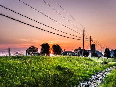 Beautiful orange sky sunrise view with a farm including a barn and silo on the horizon with a stone lined ditch and power lines in foreground above. clipart