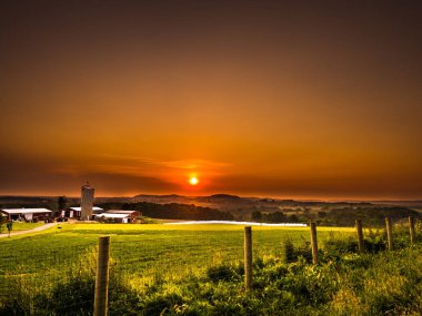Beautiful orange and yellow colorful sunset over a rural farm or country scene in Galena Illinois with wood post and wire fence in foreground and rolling hills, barn and silo beyond. clipart