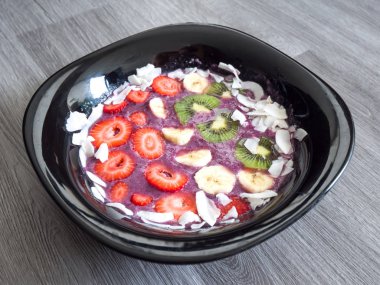 Canlı renkli ve taze meyve acai kase veya mor renkli blueberry smoothie çilek, muz, kivi güler yüzlü kase tepesinde ve cddeki arka plan üzerinde siyah bir kase astar Hindistan cevizi pul