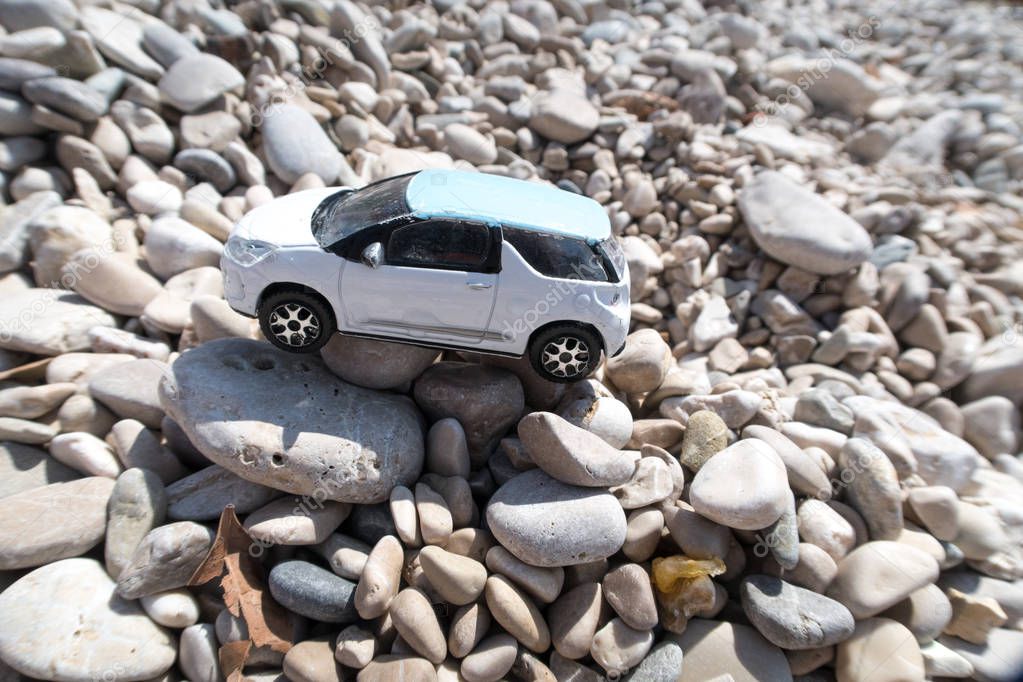 A closeup photograph of a white colored European toy car resembling a sports utility vehicle or SUV laying on a rocky beach with white rounded pebbles below.