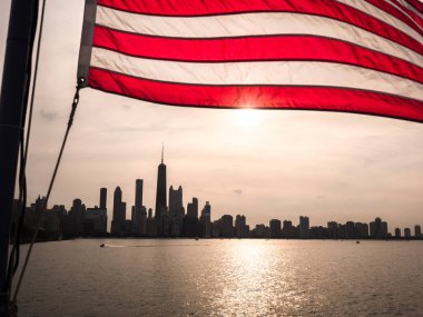 Bir Amerikan bayrağı rüzgarda üzerinde Chicago manzarası ile bina siluetleri ve Michigan Gölü su üzerinde yansıtan güneş batımında dalgalanan güzel bir resim.