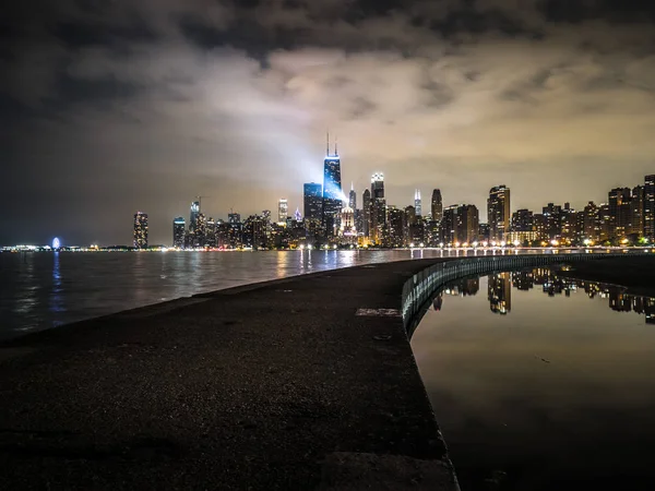 Beautiful night photograph looking down the concrete pier near North Avenue Beach in Chicago with the famous skyline reflecting on the water of Lake Michigan beyond.