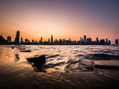 Chicago manzarası resim yukarıda mor ve turuncu gökyüzü ve dalgalar ve su Michigan Gölü ve ön planda beton kıyı dalgalanan ile siluetleri bina ile güzel bir gün batımı sırasında.