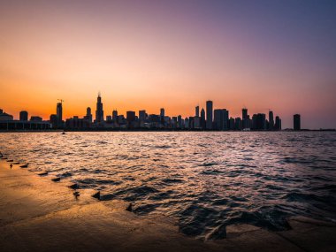 Chicago manzarası resim yukarıda mor ve turuncu gökyüzü ve dalgalar ve su Michigan Gölü ve ön planda beton kıyı dalgalanan ile siluetleri bina ile güzel bir gün batımı sırasında.