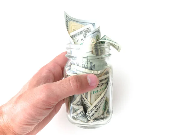 A closeup photograph of a white Caucasian male hand holding a glass mason savings jar with wads of cash hanging out of the top isolated on a white background.
