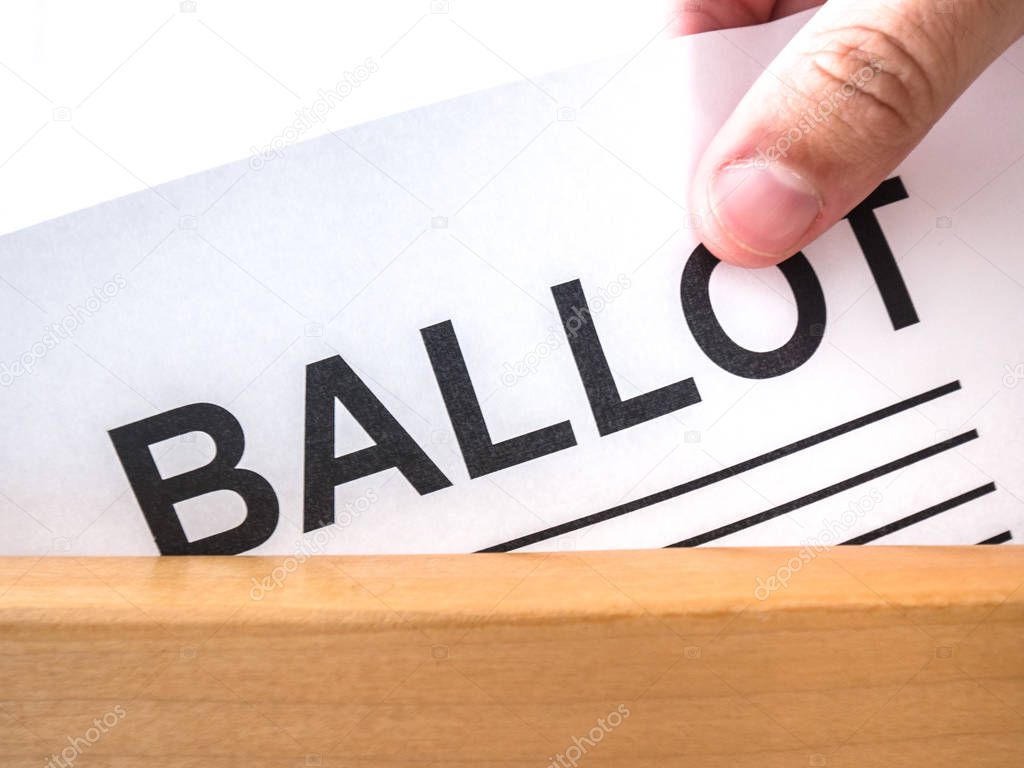 A close up photograph of a white Caucasian male hand dropping a voting ballot paper into a wooden box.