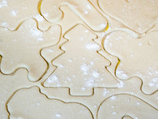 A closeup of a Christmas tree cutout and other holiday stencils or shapes including gingerbread man cut in raw sugar cookie dough with flour sprinkled on top making a beautiful festive background.