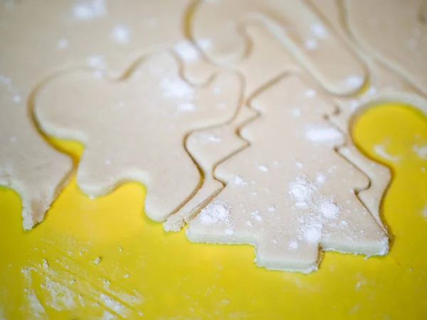 A closeup of a Christmas tree cutout and other holiday shapes in raw sugar cookie dough on a yellow cutting board with flour sprinkled on top making a festive background image for the holidays.