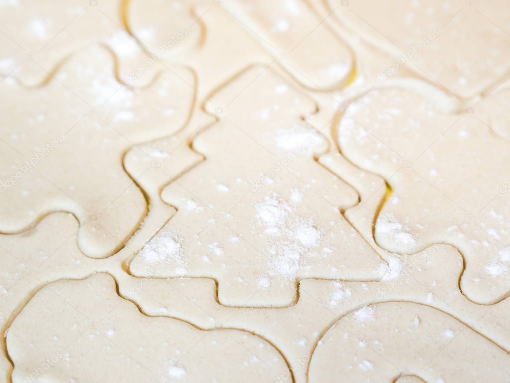 A closeup of a Christmas tree cutout and other holiday stencils or shapes including gingerbread man cut in raw sugar cookie dough with flour sprinkled on top making a beautiful festive background.