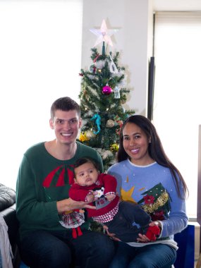 Karışık yarış çift tutun onların sevimli bebek bebek çocuk onlar kendi şenlikli kırmızı, yeşil ve mavi çirkin Noel Kazaklar ve tatil için bir ağacın önünde poz.