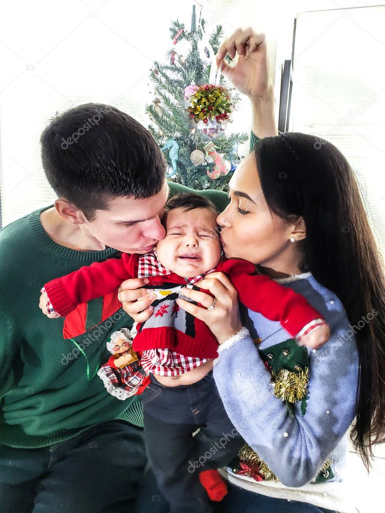 A mixed race couple of Caucasian and African American decent hold their screaming and crying baby up as they kiss his cheeks wearing festive ugly Christmas sweaters while holding a mistletoe above.